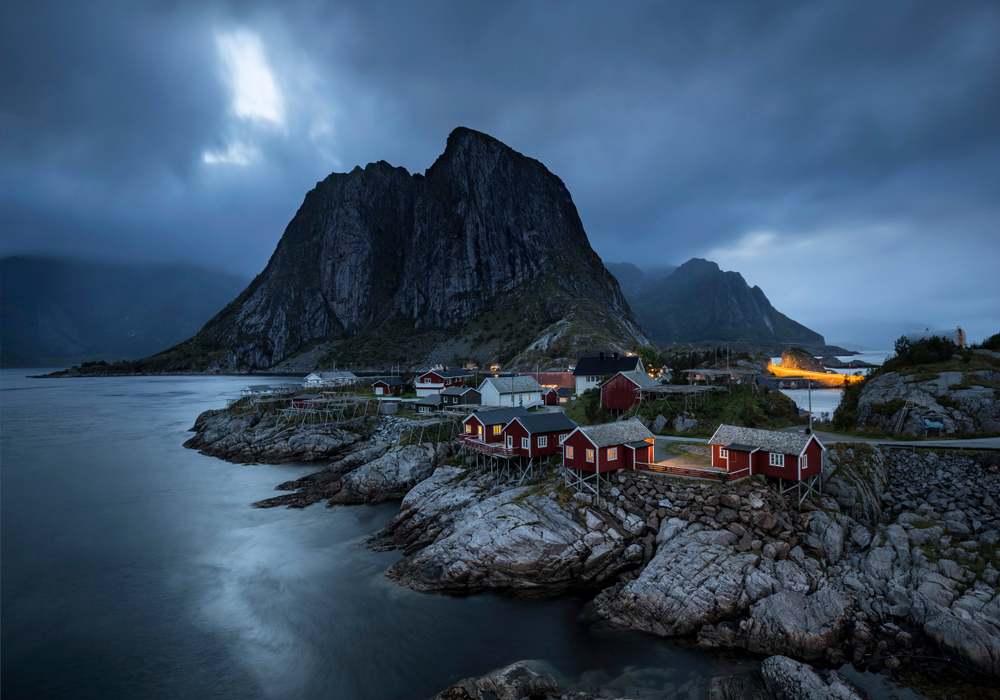 Little red buildings near the ocean by mountains Summer midnight in Hamnoy (Hamnøya in Norwegian), Lofoten. Lofoten is an archipelago in the county of Nordland, Norway, known for a distinctive scenery with dramatic clouds, mountains and peaks