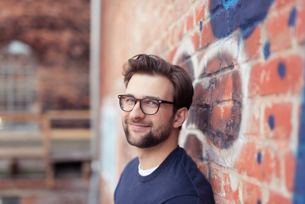 Man with Facial Hair Wearing Eyeglasses and Leaning Against Brick Wall Painted with Graffiti