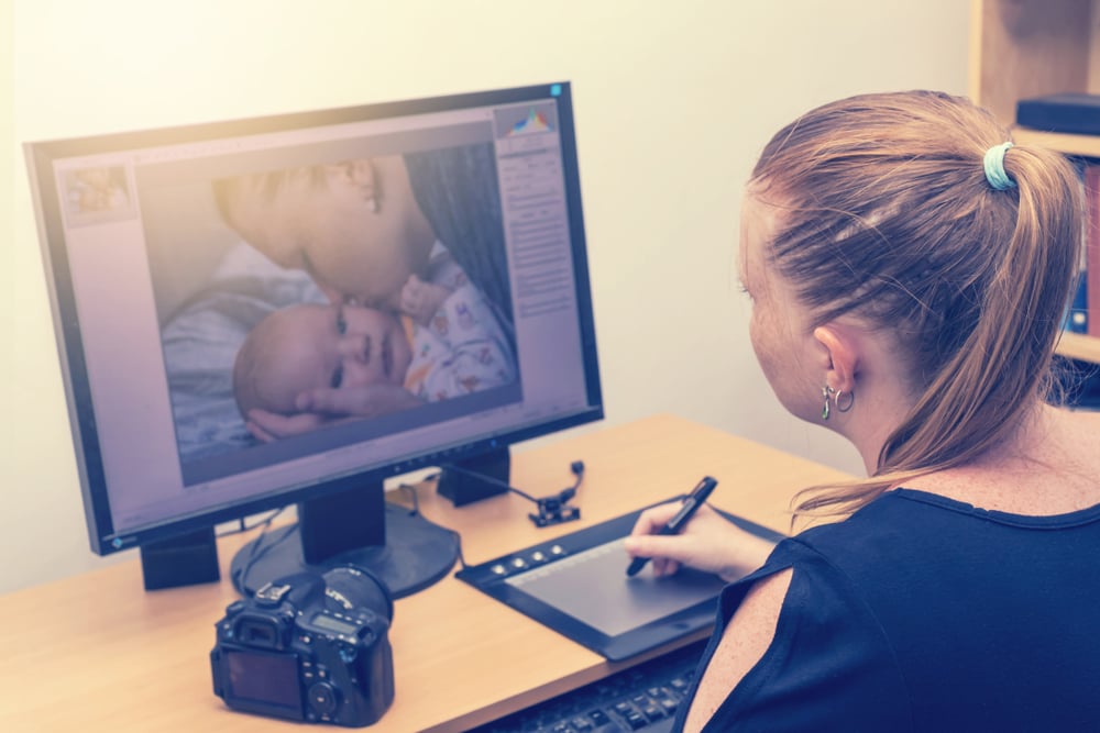 woman is editing photos by a graphics tablet and graphics software. Photo of kissing mother of her baby is on computer monitor in front of her.