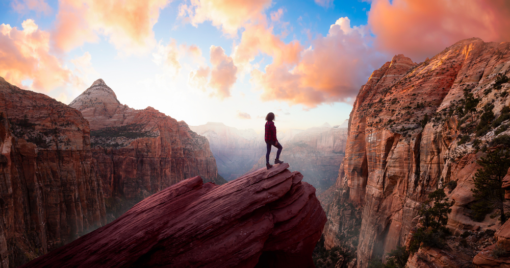 Adventurous Woman at the edge of a cliff is looking at a beautiful landscape view in the Canyon during a vibrant sunset. Taken in Zion National Park, Utah, United States. Sky Composite Panorama