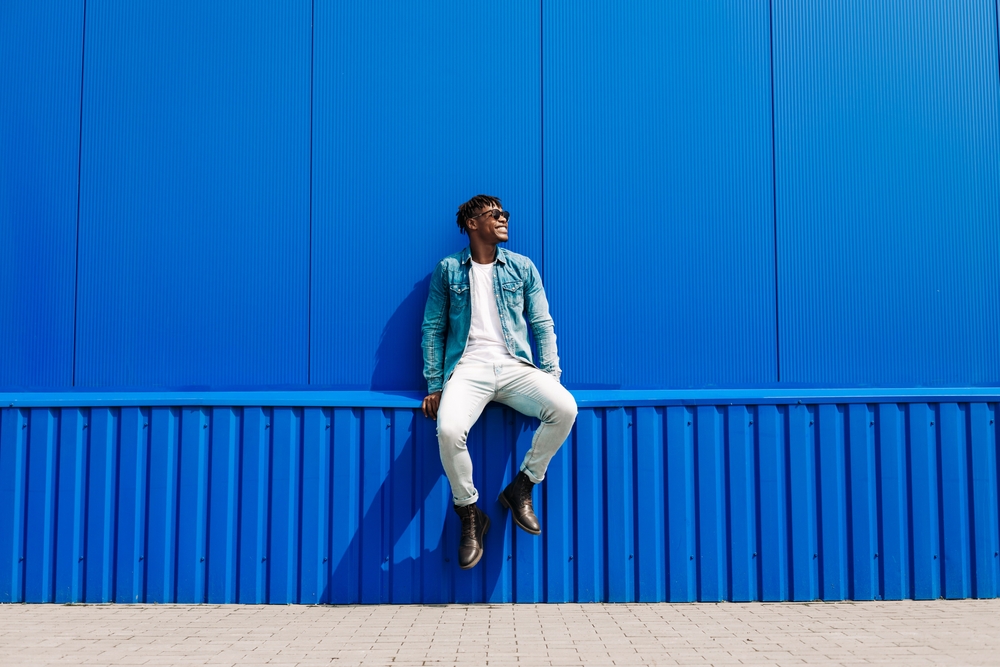 african male fashion model in sunglasses smiling and posing while standing near blue wall outdoors.