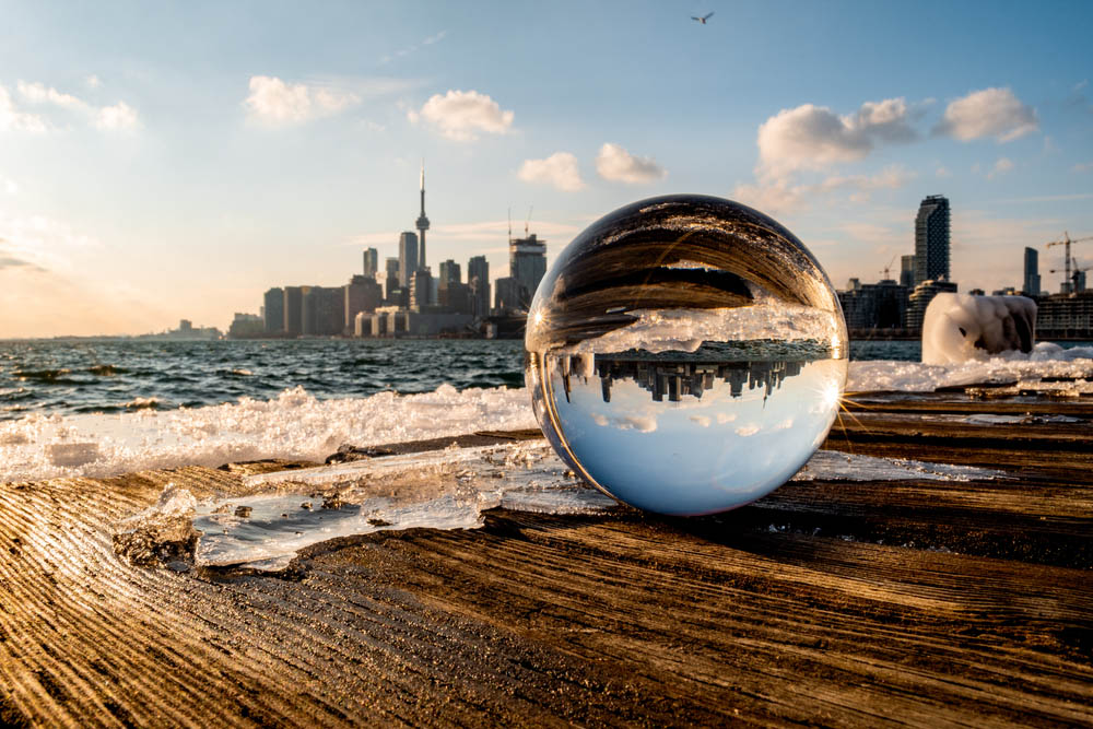 A blurry Toronto skyline behind a lensball with its reflection at Polson Pier as the sun is setting on a cold winter day.