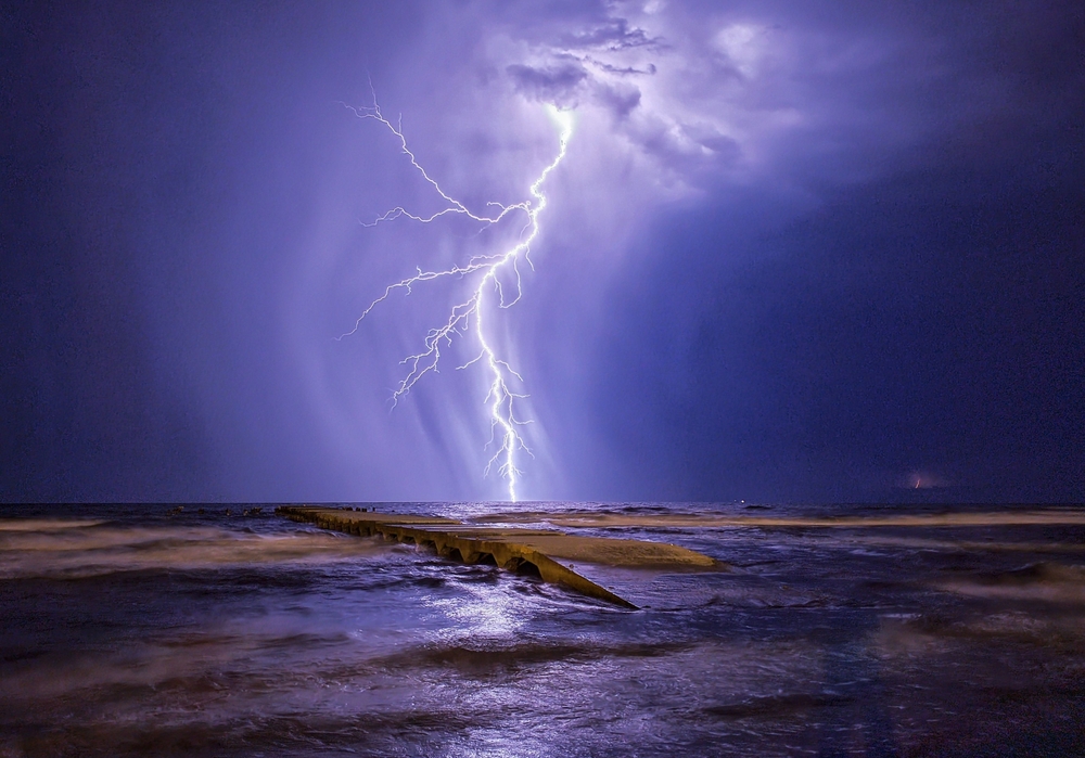 Dramatic image of a pier illuminated by a powerful bolt of lightning striking over the ocean in the background