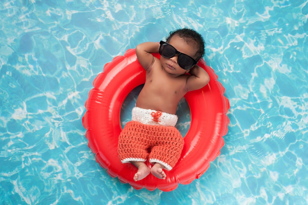 newborn baby boy sleeping on a tiny inflatable swim ring. He is wearing crocheted board shorts and black sunglasses.