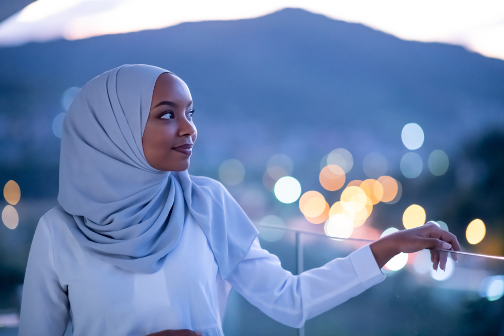 African modern Muslim woman in night at balcony smiling at camera with city bokeh lights in background