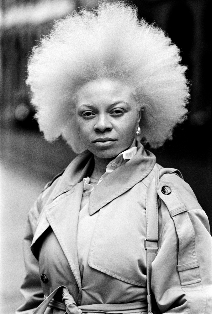 Serious female albino model showing strength in this monochrome film portrait in the street. She is wearing a trench coat and her hair is blonde and curly. Moody portrait with a lot of organic grain.